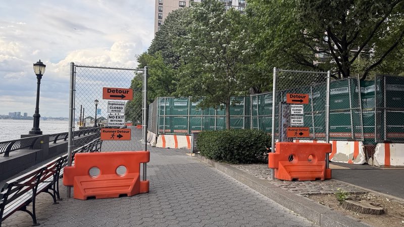 Two chain-link gates closing the esplanade at Albany Street, hung with detour and esplanade-closed signs, orange water barriers at their feet and green-screened construction fence behind