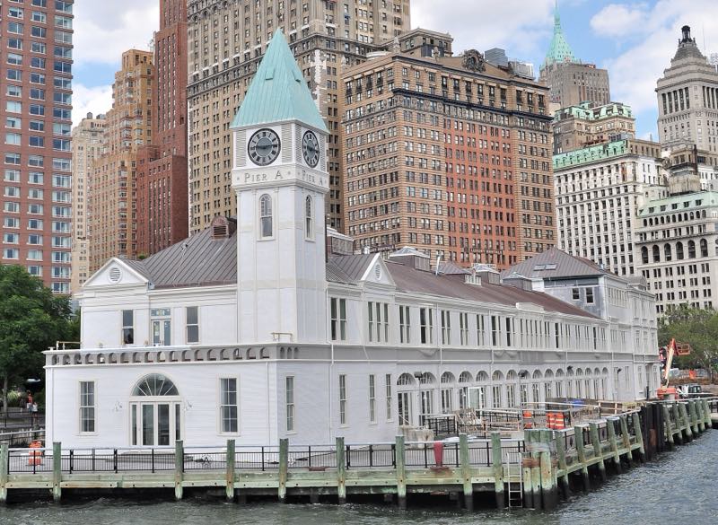 Pier A seen from the water, its white clock tower and long pier shed on timber piles, orange construction barriers on the plaza at right and Lower Manhattan's towers behind