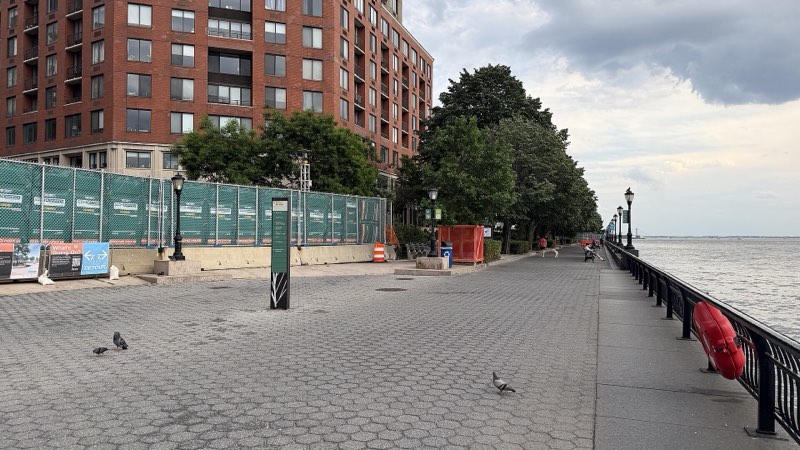 The esplanade at Rector Place, green-screened fence and detour boards along its inland side, the river railing and a life ring at right, a few people walking south