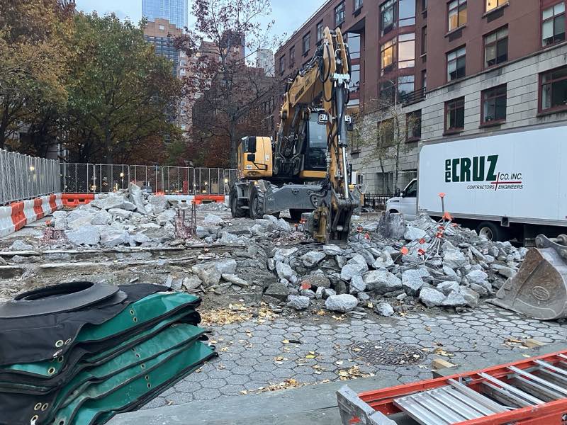 A yellow excavator in a field of broken concrete where the Upper Room stood at Albany Street, construction fencing and a contractor's truck beside it, apartment buildings behind