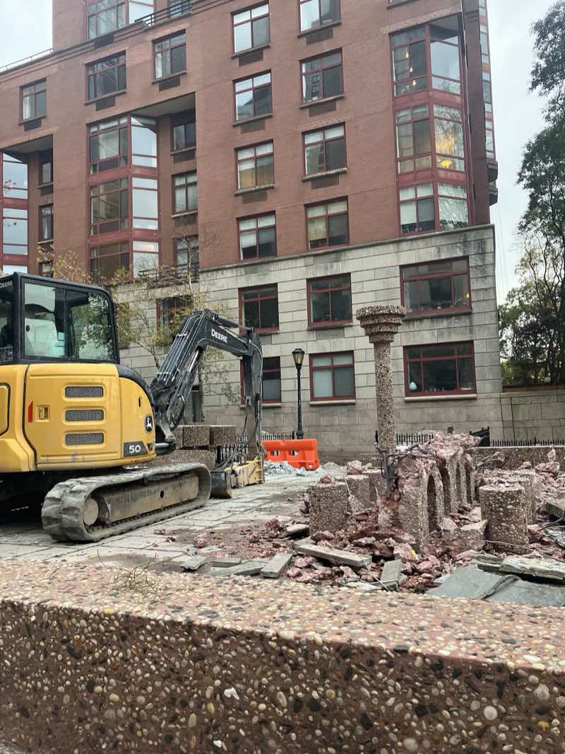 A yellow mini excavator beside the Upper Room's broken pebbled columns, one column still standing among the rubble, an apartment building behind