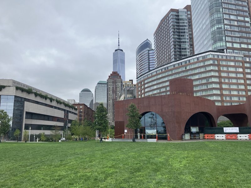 The rebuilt Wagner Park lawn under a grey sky, the new rust-red arched pavilion behind orange and white barriers and a Pardon Our Appearance sign, the Museum of Jewish Heritage at left and One World Trade Center beyond