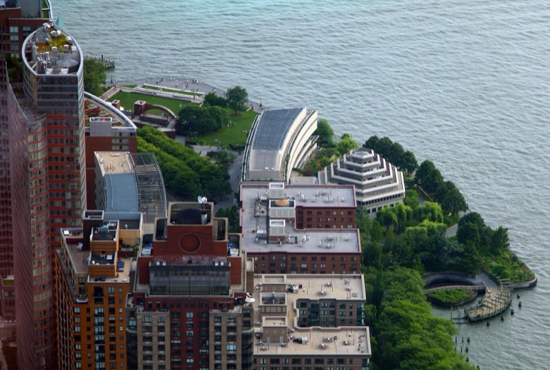 Wagner Park from high above in June 2015: the old brick pavilion and its lawn at the water's edge, mature trees along the shore, the stepped Museum of Jewish Heritage, and Battery Park City's apartment blocks in the foreground